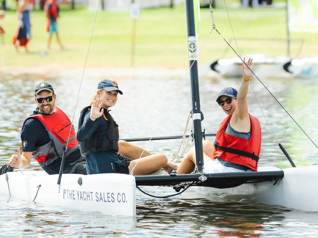Three adults in lifejackets smiling and waving from a small catamaran on Oxenford lake